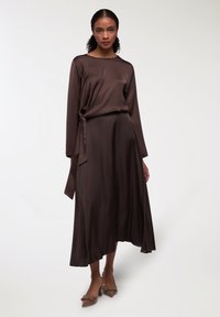 Abito lungo in satin marrone con maniche lunghe e dettaglio in vita annodato, che presenta una gonna A-line fluttuante. Texture liscia e silhouette elegante. Tacchi a punta marroni.