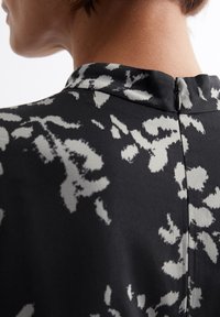 Svart bluse med et subtilt grått blomstermønster, med høy nakke og glidelås i ryggen. Glatt stofftekstur.