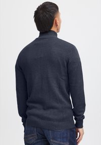 Un pull en maille noir gris, avec un col roulé, présentant des poignets et un ourlet côtelés, vu de dos sur fond blanc.