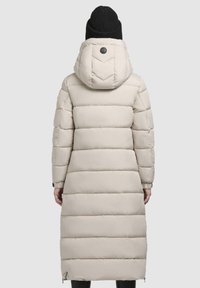 Beige gevoerd lang jack met een capuchon, voorzien van chevronstiksels, ritszakken aan de zijkant en geribbelde manchetten. Zwarte gebreide beanie op het hoofd.