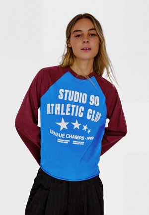 Vrouw met lang blond haar draagt een blauw en bordeauxkleurig shirt met lange mouwen met tekst en sterren "STUDIO 90 ATHLETIC CLUB LEAGUE CHAMPS 1999".