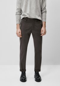 Homme portant un pull gris, un pantalon brun foncé à revers et des chaussures en cuir noires, se tenant contre un fond gris clair uni.