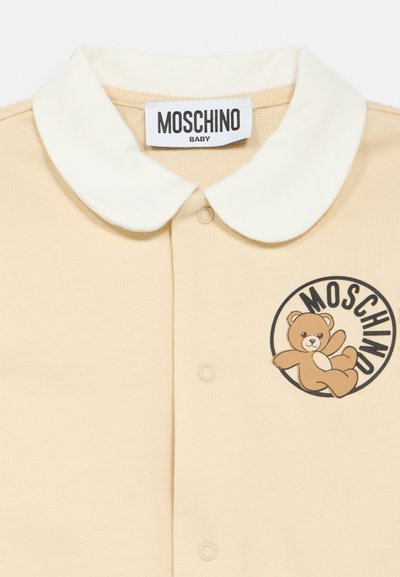 Πόλο μπλούζα σε ανοιχτό μπεζ βαμβάκι με κρεμ γιακά, με καφέ cartoon αρκουδάκι λογότυπο και στρογγυλό κείμενο "MOSCHINO" στο στήθος.