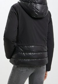 Persona che indossa una giacca a vento nera con cappuccio, con un retro imbottito lucido e maniche, abbinata a jeans di denim grigi.