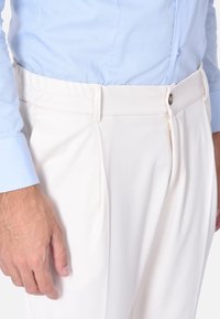 Pantaloni bianchi con una vestibilità su misura, caratterizzati da una parte frontale piatta, tasche laterali e una chiusura con zip. Il tessuto sembra liscio con una leggera lucentezza.