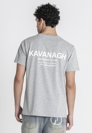 Jonge man met krullend haar die een grijze T-shirt draagt met witte tekst "KAVANAGH WINNERS CLUB gegenereerd door mensen voor kampioenen" op de rug.