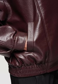 Mano con pantalones negros metida en el bolsillo de una chaqueta de cuero texturizada de color burdeos oscuro con dobladillo elástico fruncido.