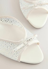 Chaussures de ballet blanches à bout ouvert avec un mesh transparent décoré de petits strass argentés et un petit nœud en tissu à l'avant.