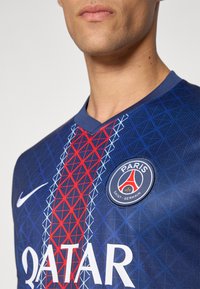 Le maillot de football Nike présente un fond bleu avec un motif géométrique rouge et un logo rond indiquant "PARIS SAINT-GERMAIN."