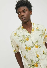 Camisa de manga corta en crema claro con un patrón de hojas verdes y naranjas amarillas por toda la prenda, que presenta un cuello clásico y una tapeta de botones.