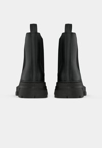 Pár čiernych členkových botas s hrubou podrážkou a ťahacími pútkami, zobrazené zozadu na jednoduchej bielej ploche.
