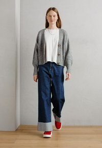 Grauer gerippter Cardigan mit drei Knöpfen über einem weißen Oberteil, kombiniert mit weit geschnittenen Jeanshosen mit grauem Saum und roten Sneakers.