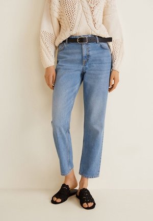 Kobieta w jasnoniebieskich dżinsach z wysokim stanem i czarnym paskiem, kremowym dzianinowym swetrze oraz czarnych, szydełkowanych sandałach na stopę bez palców na wsuwce.