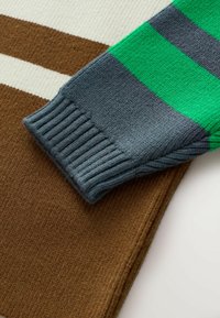 Manche de pull en tricot gris, ornée de rayures vertes, reposant sur un tissu texturé marron. Détail de poignet côtelé visible.