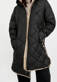 Sort quiltet dunjakke med hætte, med beige foring og detaljer, lynlåslukning og trykknapper langs siderne.