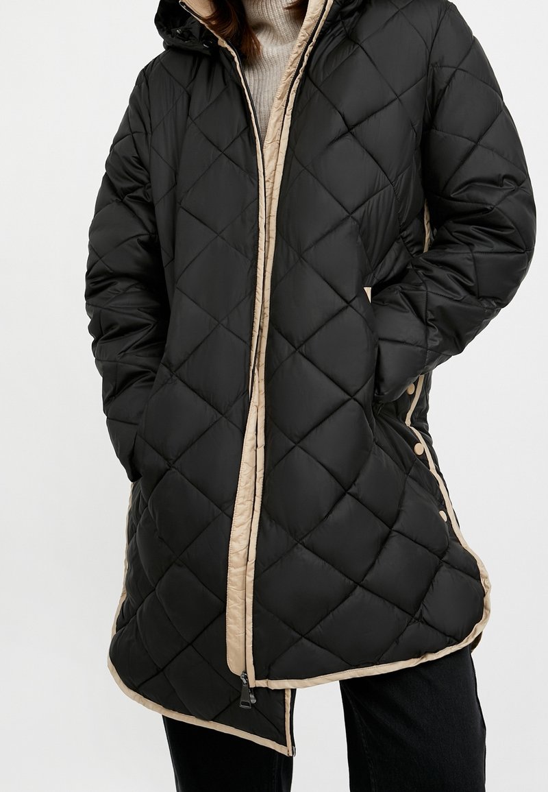 Sort quiltet dunjakke med hætte, med beige foring og detaljer, lynlåslukning og trykknapper langs siderne.
