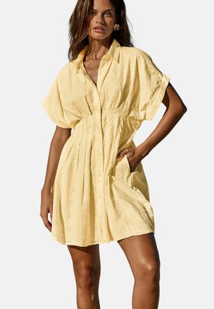 Frau trägt ein gelbes Lochstickerkleid mit kurzen Ärmeln und Knöpfen, posiert mit einer Hand in der Tasche vor einem weißen Hintergrund.