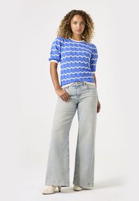 Camisa de punto azul con textura y patrón de zigzag blanco, combinada con jeans de pierna ancha de lavado claro y zapatos de tacón crema.