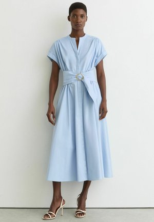 Vrouw draagt een lichtblauwe midi-jurk met korte mouwen en een brede riem met een ronde gesp, gecombineerd met beige sandalen met hoge hakken.