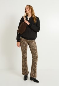 Chaqueta bomber negra acolchada, suéter crema, pantalones acampanados marrón con estampado de leopardo, y botines negros de tacón bloque. Llevando un bolso marrón texturizado.
