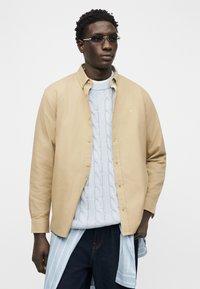 Νέος άνδρας με σκούρο δέρμα, φοράει φωτισμένα γυαλιά, μπεζ πουκάμισο με κουμπιά πάνω από μπλε πλεκτό πουλόβερ, ανοιχτό ριγέ πουκάμισο δεμένο γύρω από τη μέση, σκούρο παντελόνι.