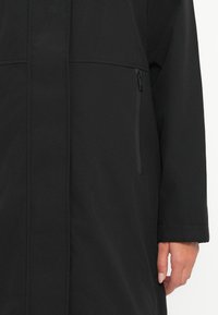 Black, long outerwear featuring a smooth texture, minimalistic design, and zippered pocket detail on the left side.