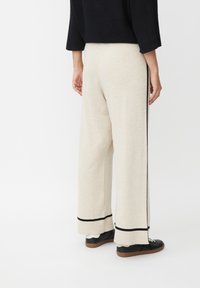 Weit geschnittene Hosen aus leichtem beigen Strickstoff mit schwarzen Seitenstreifen und gerippten Säumen, kombiniert mit schwarzen Schuhen.