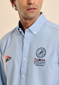 Camisa abotonada de color azul claro con cuello. Presenta logotipos bordados, incluyendo un velero y banderas náuticas. Tejido de algodón, textura suave.