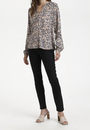 Kvinde iført en langærmet mønstret beige bluse, sorte tætsiddende bukser og lyse beige åbne hælede mule-sko, stående foran en hvid baggrund.