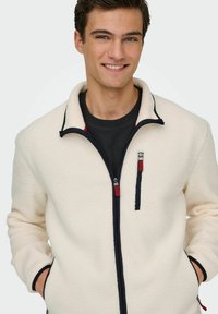 Lächelnder Mann in einer cremefarbenen Fleecejacke mit schwarzen Reißverschlussdetails und roten Reißverschlusszügen, die Hände in den Taschen, vor einem schlichten Hintergrund.