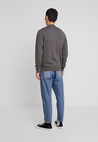 Mann, der von hinten steht, trägt ein graues langärmliges Shirt, blue Jeans und schwarze High-Top-Sneaker vor einem schlichten hellen Hintergrund.