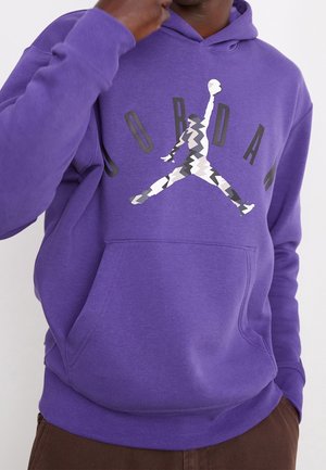 Osoba ubrana w fioletową bluzę z kapturem ze stylizowanym logo Jumpman i napisem "JORDAN" na piersi, ręka przy twarzy, widoczne brązowe spodnie.