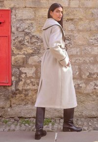 Palton gri deschis cu un guler lat, accente negre, buzunare laterale și o curea, combinat cu cizme negre până la genunchi, sprijinit de un zid de piatră.