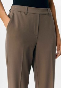 Pantalons marron ajustés avec passants de ceinture, poches latérales et un pli à l'avant, portés avec un haut noir rentré, la main reposant dans la poche.
