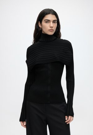 Femme aux longs cheveux foncés portant un pull noir côtelé boutonné avec un col haut asymétrique et un pantalon noir sur un fond uni.