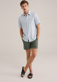 Világoskék, rövid ujjú ing gombos elülső résszel; zöld rövidnadrág; fekete slip-on szandál; a modell keze a zsebében áll, semleges háttérben.