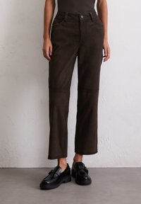 Braune Wildlederhosen mit geradem Schnitt, ausgestattet mit einem Knopfverschluss und Fronttaschen. Kombiniert mit schwarzen Slippern für einen Kontrast.