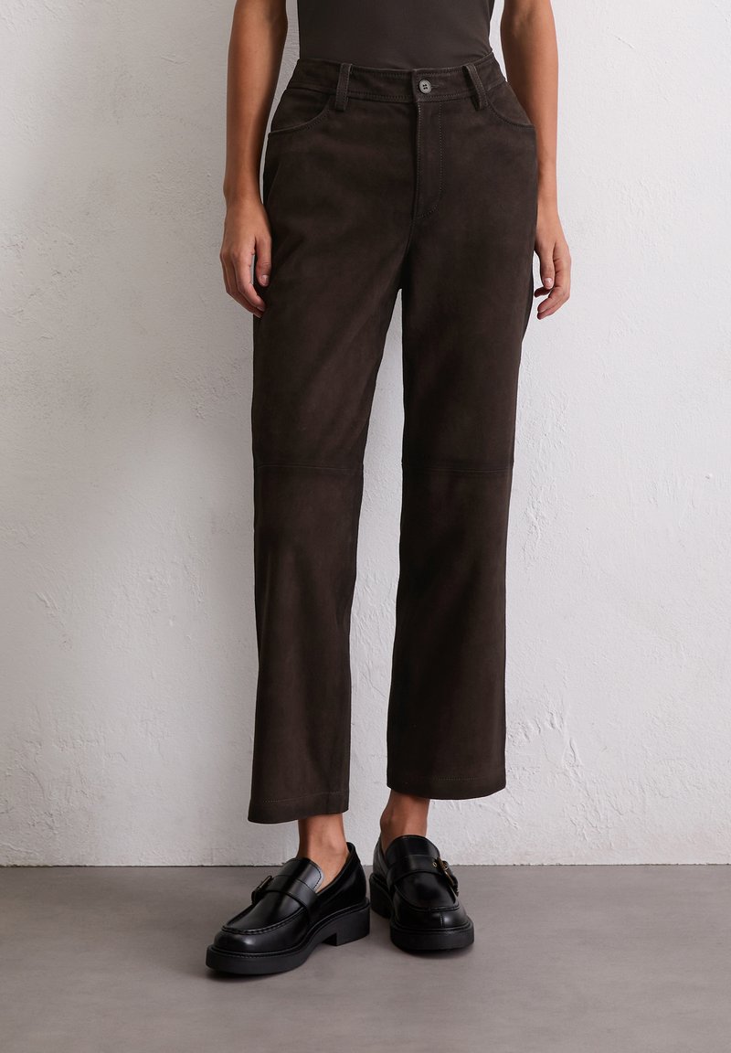 Braune Wildlederhosen mit geradem Schnitt, ausgestattet mit einem Knopfverschluss und Fronttaschen. Kombiniert mit schwarzen Slippern für einen Kontrast.