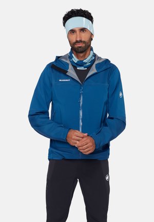 Mand iført en blå Mammut-jakke, sorte bukser, lyseblåt pandebånd og et blåt mønstret halsrør mod en ensfarvet baggrund.
