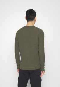 Chemise manches longues vert olive avec encolure ronde et ourlet cousu, associée à un pantalon cargo foncé. Le tissu semble doux et texturé.