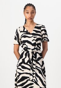 Svart og kremfarga zebra-striped innpakningskjole med korte ermer, v-hals og bånd i livet. Glatt stoff med en flytende design.