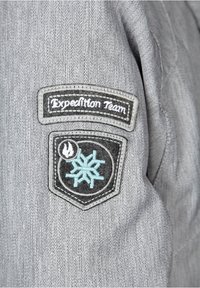 Grå tyglapp med fiskbensmönster som har två broderade märken; "Expeditionsteam" i vitt på en mörk bakgrund och en blå snöflinga på svart.