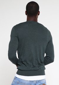 Mężczyzna w ciemnozielonym swetrze z długim rękawem i jasnoniebieskich dżinsach, pokazany z tyłu, stojący na jednolitym białym tle.