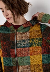 Jonge vrouw in een patchwork gebreide zip-up trui met groene, gele, rode en bruine tinten, die met haar linkerhand de capuchon aanpast.