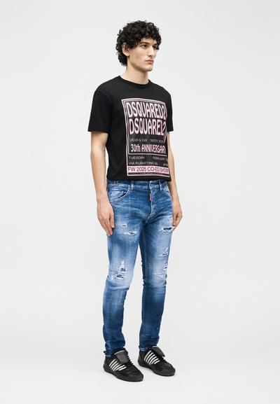 Άνδρας με σγουρά μαλλιά που φοράει μαύρο μπλουζάκι DSQUARED2 με σχέδιο, μπλε στενά τζιν με φθορές και μαύρα αθλητικά παπούτσια με λευκές ρίγες, όρθιος.