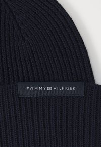 Jūros mėlynumo megzta kepurė su raštuotu paviršiumi, kuriame yra stačiakampio formos juodas etiketė, išraidžių "TOMMY HILFIGER" baltais simboliais.