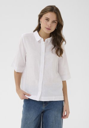 Femme aux longs cheveux bruns portant des boucles d'oreilles en perles, une chemise blanche à manches courtes boutonnée et un jean bleu, debout avec une main dans la poche.