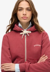 Sudadera roja con cremallera y capucha ajustable, de tejido suave, con bolsillos frontales y el logotipo blanco "SUPERDRY" en el pecho, llevada sobre una camiseta a rayas.