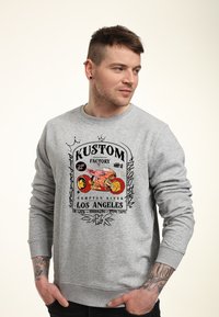 Uomo con orecchini e tatuaggi che indossa un felpa grigia con una grafica di una moto rossa e il testo "Kustom Factory Los Angeles", che guarda a destra.
