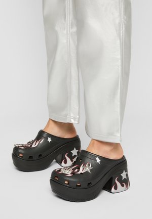 Sabots à plateforme noirs avec des motifs de flammes et d'étoiles portés avec un pantalon blanc, montrant les chevilles et les pieds sur un fond clair.